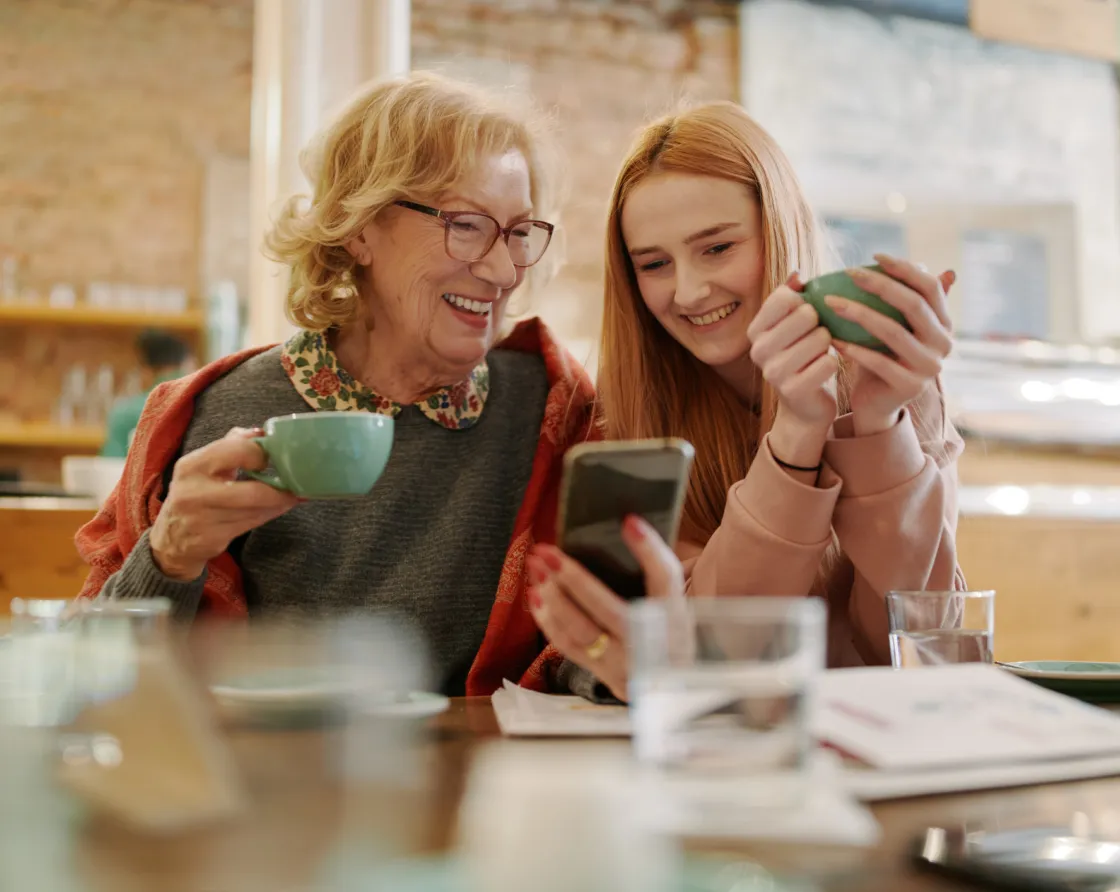 Foto Großmutter und Enkeltochter die bei einer Tasse Kaffee lächelnd ein Mobiltelefon ansehen.